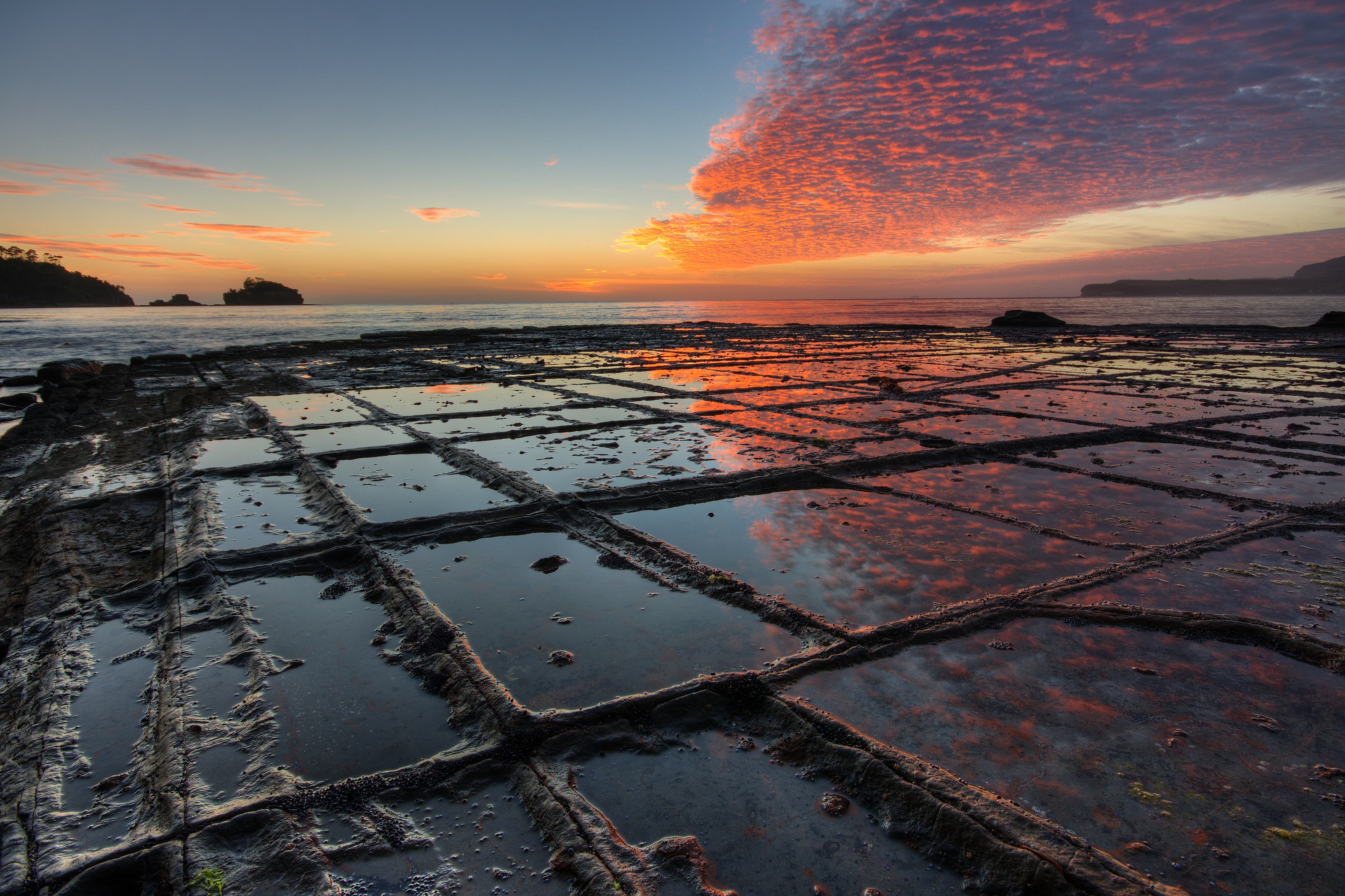 Sunrise, “Tessellated Pavement”, Eaglehawk Neck, Tasman Peninsula, Tasmania, Australia. The „Tesselated Pavement“ is the result of an orthogonal joint pattern in the rock. On the picture it shows the so called “pan formation”, where the rock in the immediate vicinity of the joints is more resistant to erosion than the rock that is more distant to the joints, This is due to alterations of the rock along the joints by hydrothermal (or similar) solutions when the rock was still buried deeply below the surface millions of years ago. When no alterations or alterations that lower the erosional resistivity have taken place in the geological past, the rock along the joints will erode faster than the rock that is more distant to the joints. In that case the so called “loaf formation” of “Tessellated Pavement” will form. by JJ Harrison